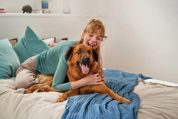 Eine lachende Person umarmt einen Hund auf einem Bett.